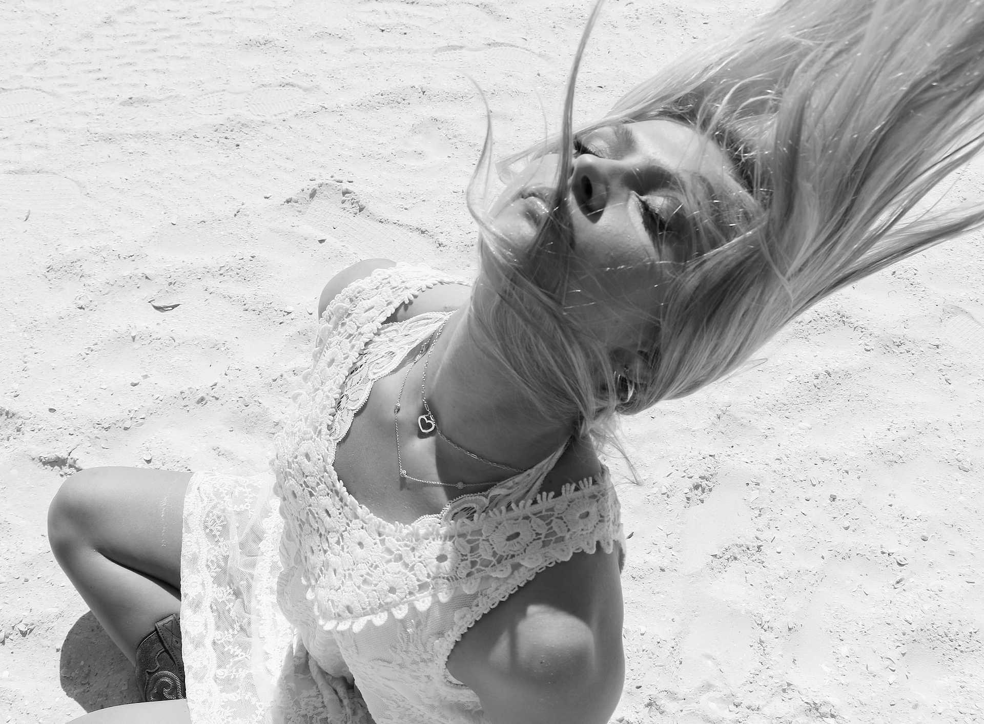 Woman in white dress with hair flowing back, sitting on sandy beach, eyes closed. Black and white.